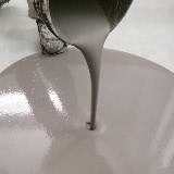 Smooth gray self-leveling floor compound pouring from a container onto a white surface, demonstrating its fluid consistency and spreading properties for interior concrete subfloors and engineered flooring applications.