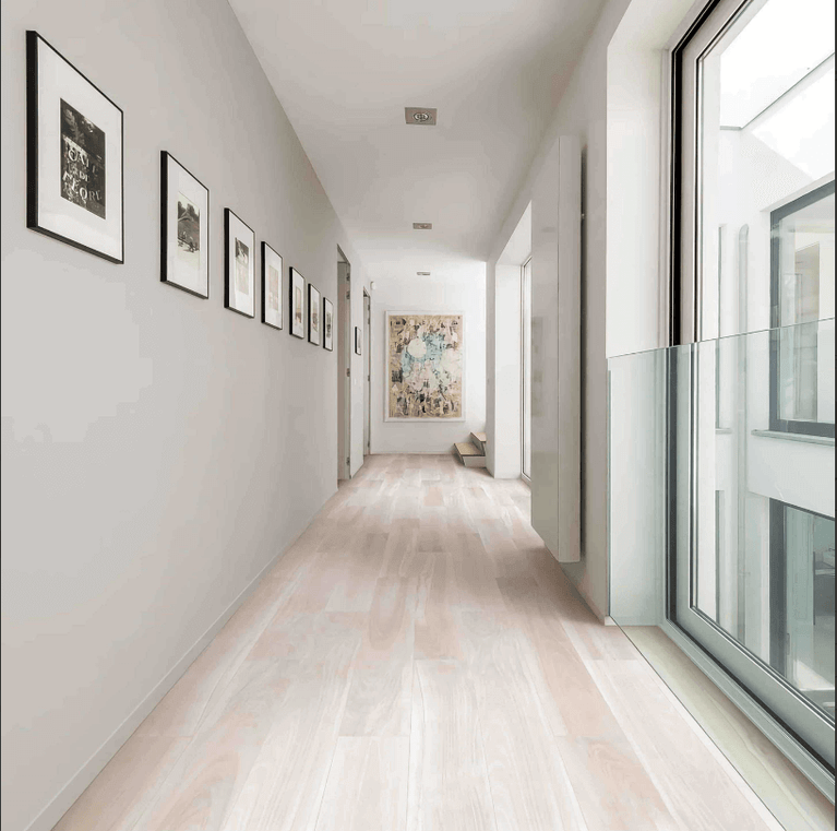 Couloir lumineux avec sol en céramique effet bois naturel Céramique Wood K Naturel, murs blancs, fenêtres et cadres décoratifs, illustrant l'élégance et la polyvalence du produit pour usage résidentiel et commercial