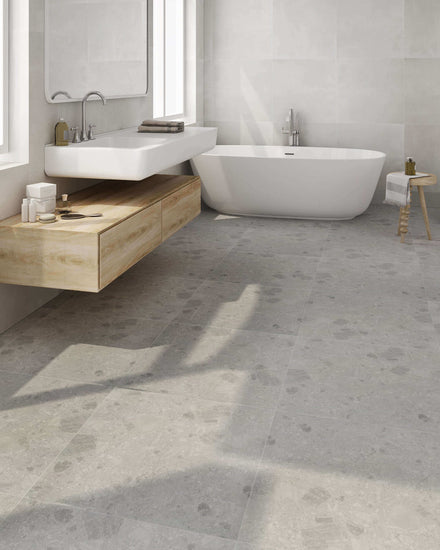 Salle de bain moderne avec carrelage gris Céramique Dolce di Grès Grey au sol, baignoire blanche autoportante, meuble vasque en bois clair suspendu et lavabo blanc, offrant une ambiance épurée et élégante.