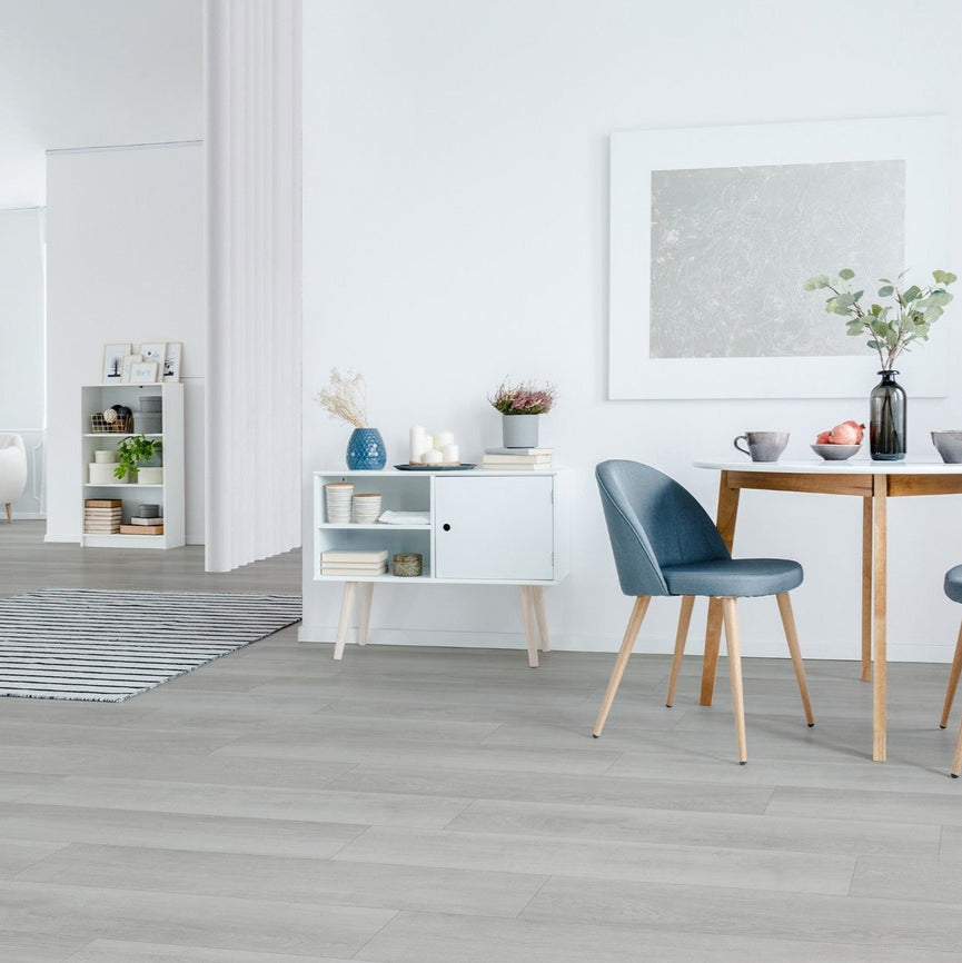 Intérieur moderne avec plancher de vinyle gris clair, table en bois, chaise bleue et meubles blancs, illustrant l'élégance et la polyvalence du revêtement Sabre pour diverses pièces. Nouveautés, En stock, Tous produits, Vinyles collés