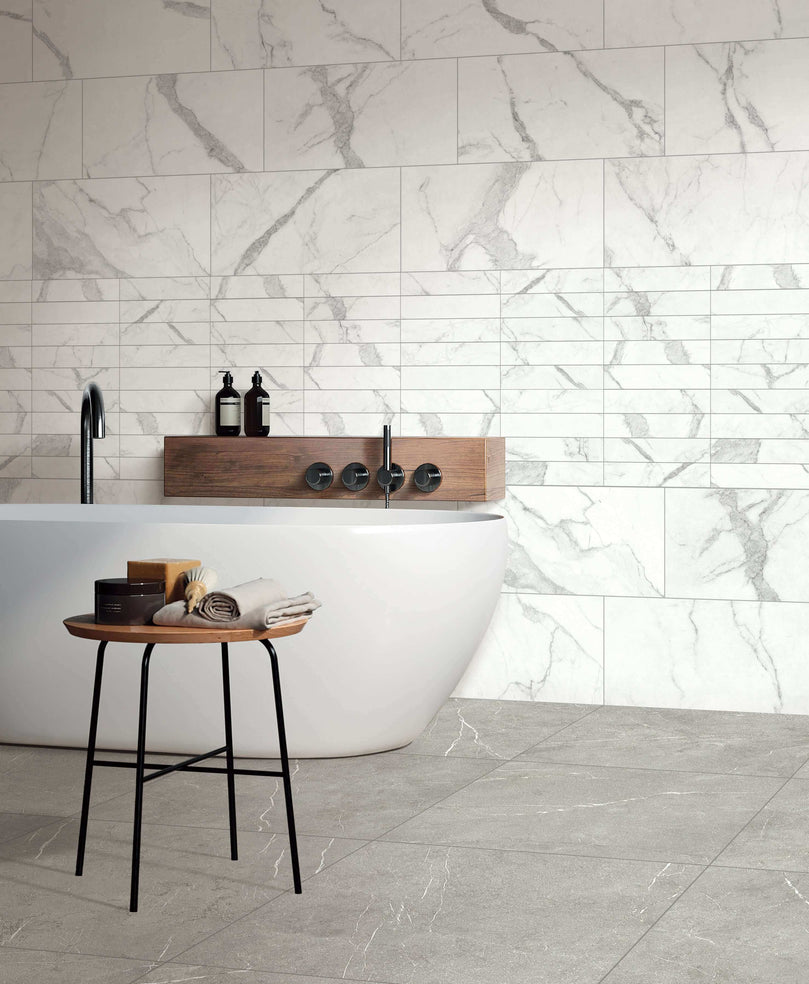 Salle de bain élégante avec carrelage en marbre Céramique Marble Stone Grigio, baignoire moderne, et table d'appoint en bois. Mur et sol en carreaux gris clair veinés, créant une ambiance luxueuse et épurée.